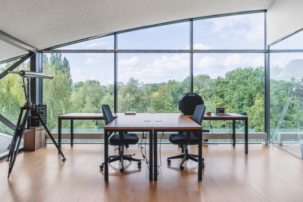 Modern kantoor met grote glazen gevel, houten vloeren, twee bureaus en een telescoop, gelegen aan de Prof. W.H. Keesomlaan met uitzicht op groen.